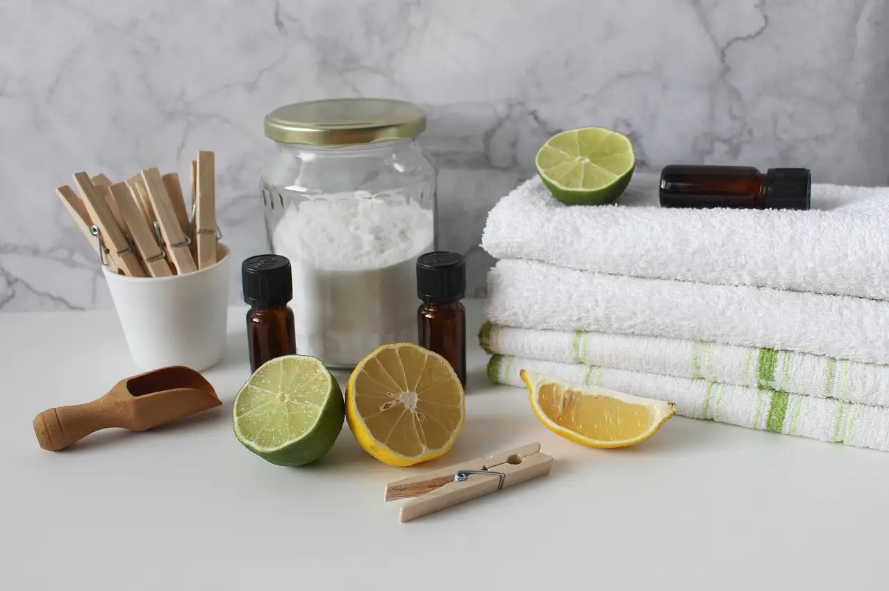 Spa setup with white towels, citrus halves, essential oil bottles, a jar of bath salts, a wooden scoop, and clothespins on a marble surface.