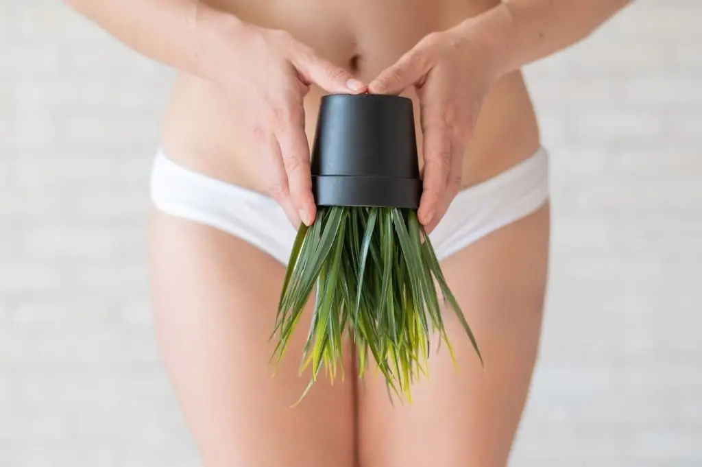 Person in white underwear holds a black handheld body brush with green palm leaf bristles in front of a light background.