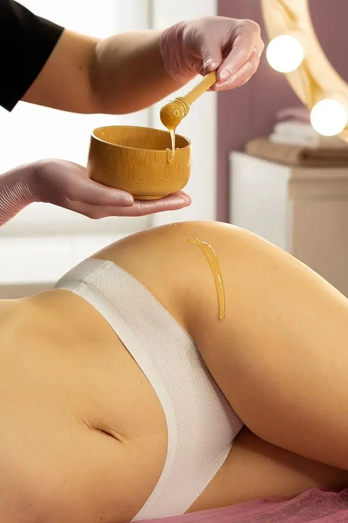 Spa practitioner drizzles honey onto a client's buttocks during a body treatment, using a wooden bowl and spatula.