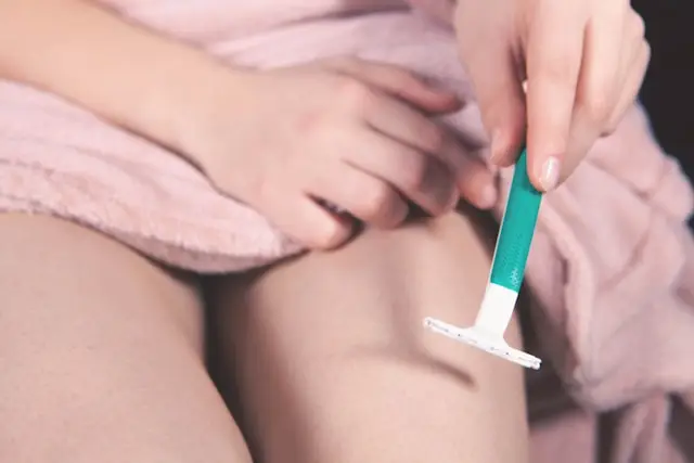 Person shaving their leg with a turquoise razor while sitting on a pink blanket bed cover.