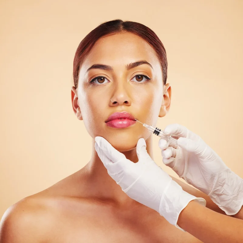 Close-up of a woman receiving a cosmetic facial injection, gloved hands hold a syringe near her mouth against a peach background.