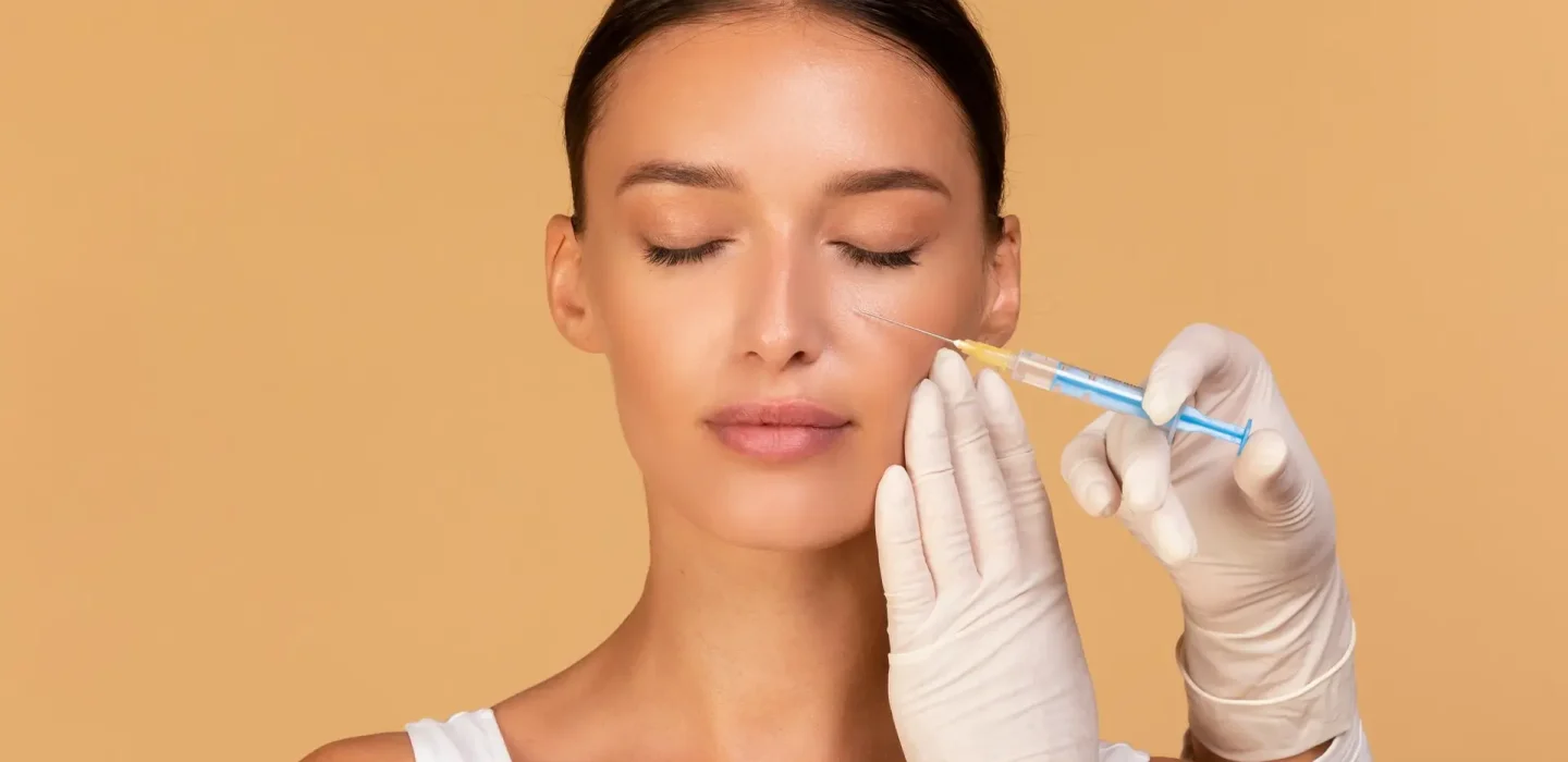 Woman with eyes closed as a clinician injects her cheek with a syringe for a cosmetic facial treatment, wearing gloves against a peach background.